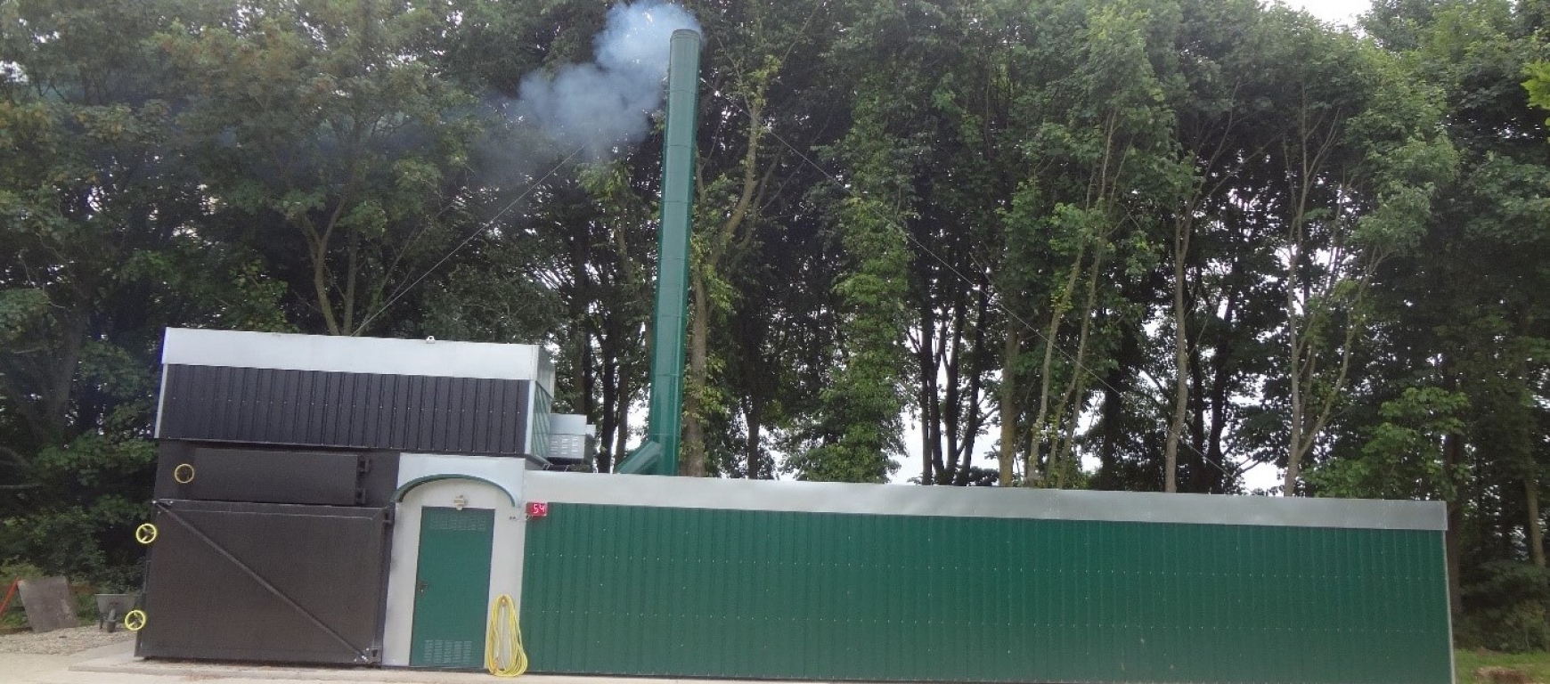 A green boiler house with a smoking chimney with trees behind